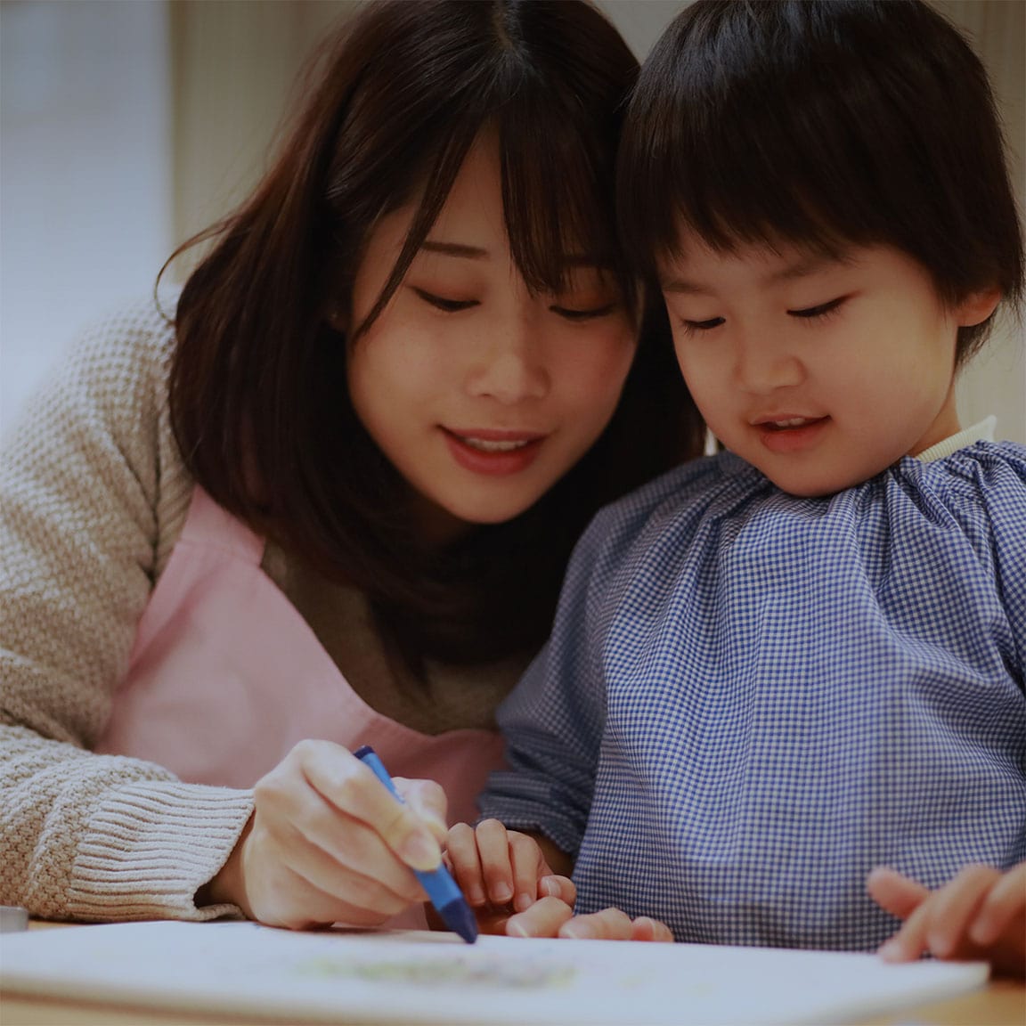 保育士の女性が子どもと一緒に机で絵を描きながら微笑んでいる場面。地域福祉や保育サービスの支え合いを象徴